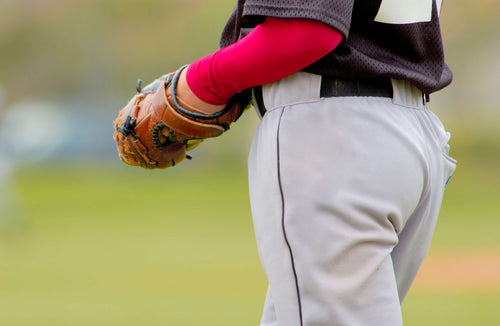 A boy wearing a baseball uniform and a red arm sleeve holds a brown baseball glove.
