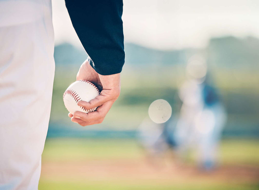 Close-up of a young pitcher's hand holding a baseball on a baseball field. A hitter and a catcher are visible in the background.