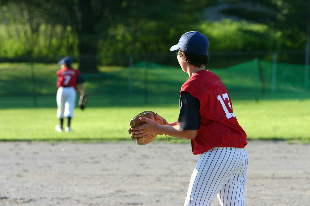 A young boy wearing a red baseball jersey with a white number "13" printed on the back, black-and-white pinstripe pants, and a blue baseball cap stands on a baseball field and holds a baseball gloves. A second baseball player is visible in the background.