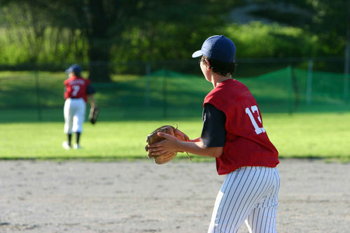 A young boy wearing a red baseball jersey with a white number "13" printed on the back, black-and-white pinstripe pants, and a blue baseball cap stands on a baseball field and holds a baseball gloves. A second baseball player is visible in the background.